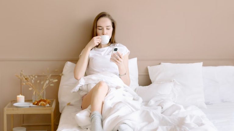 Relaxed woman in bed drinking coffee and using smartphone in a cozy setting.